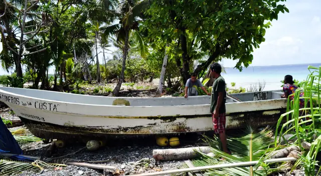 Two men work on the Roneros de la Costa beached on a Pacific shoreline. Logs beneath the hull are a centuries-old island technique for rolling vessels across sand without machinery.